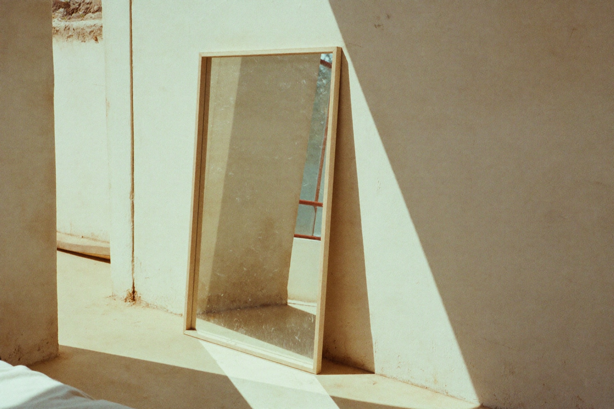A framed mirror propped against a sunlit plaster wall, reflecting only the empty room
